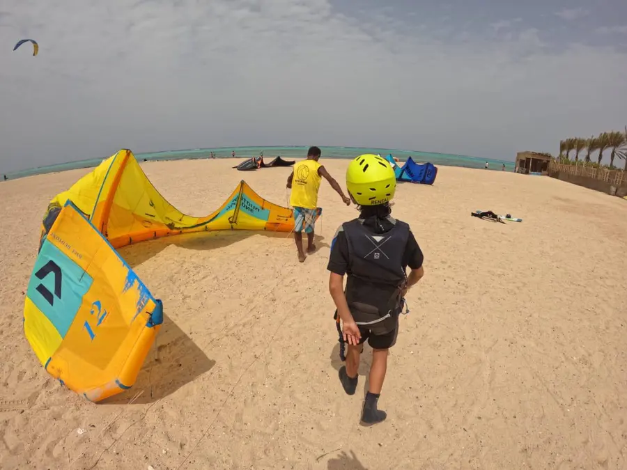Young kiter in a yellow helmet, vest and harness follows an instructor in a yellow bib across the sand to a yellow kite